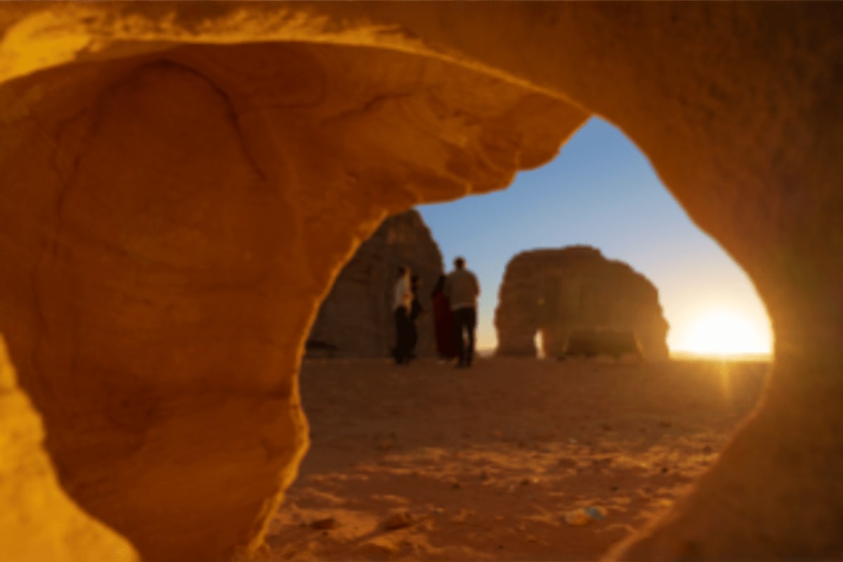 View through natural rock arch opening to tourists and desert landscape at sunset in Saudi Arabia