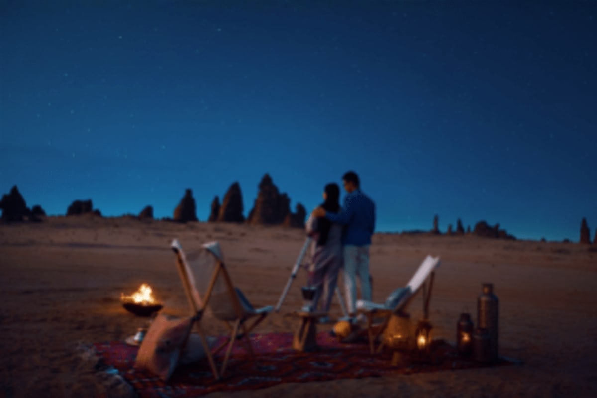People gathered around a beach bonfire at night with rock formations silhouetted against a starry sky in Saudi Arabia
