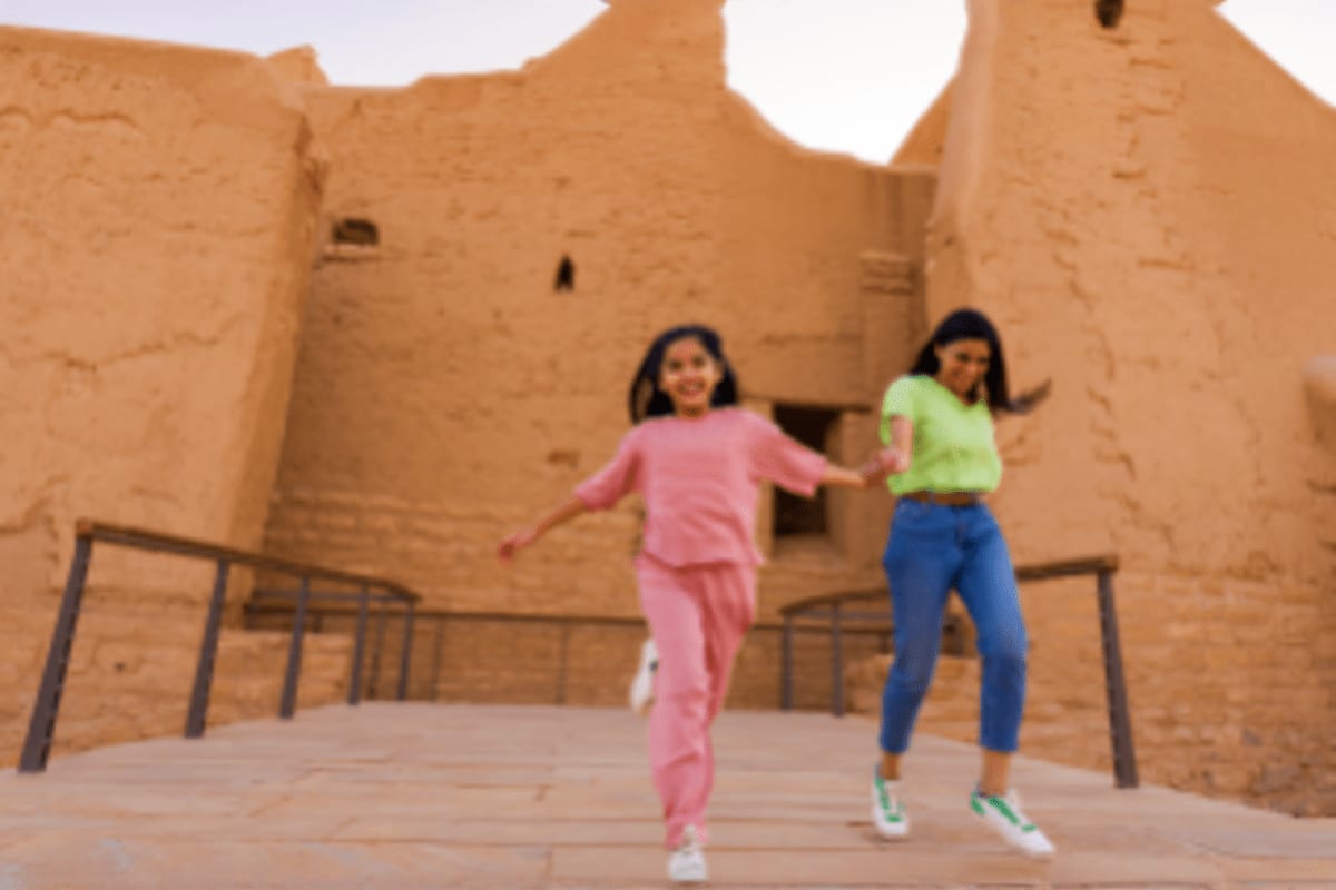 Two girls running and playing in front of ancient mud-brick fortress walls under bright sunlight in Saudi Arabia
