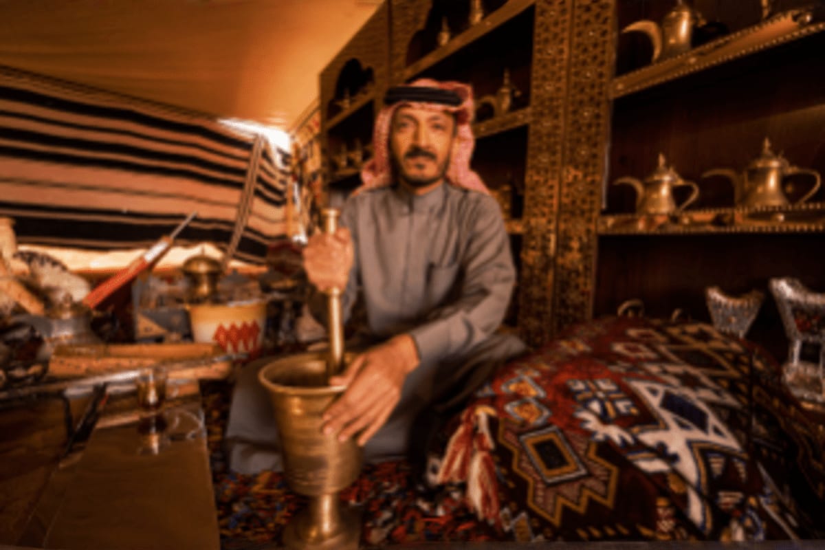 Man in traditional Saudi dress sitting in ornate tent displaying brass coffee pots and decorative vessels on patterned rugs