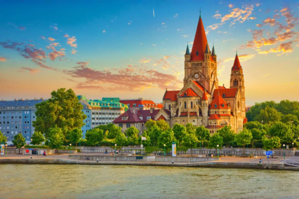 Vienna cityscape at sunset featuring St. Leopold Church with red spires along the Danube River and historic buildings