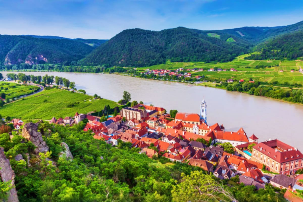 Aerial view of Dürnstein village with red-roofed houses along the Danube River, surrounded by green vineyards and forested hi
