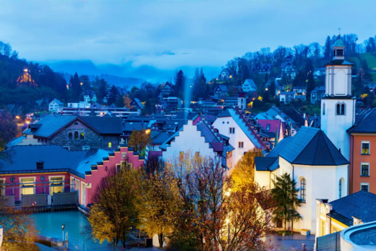 Twilight view of an Alpine village with colorful buildings, a white church with steeple, and forested mountains in the backgr