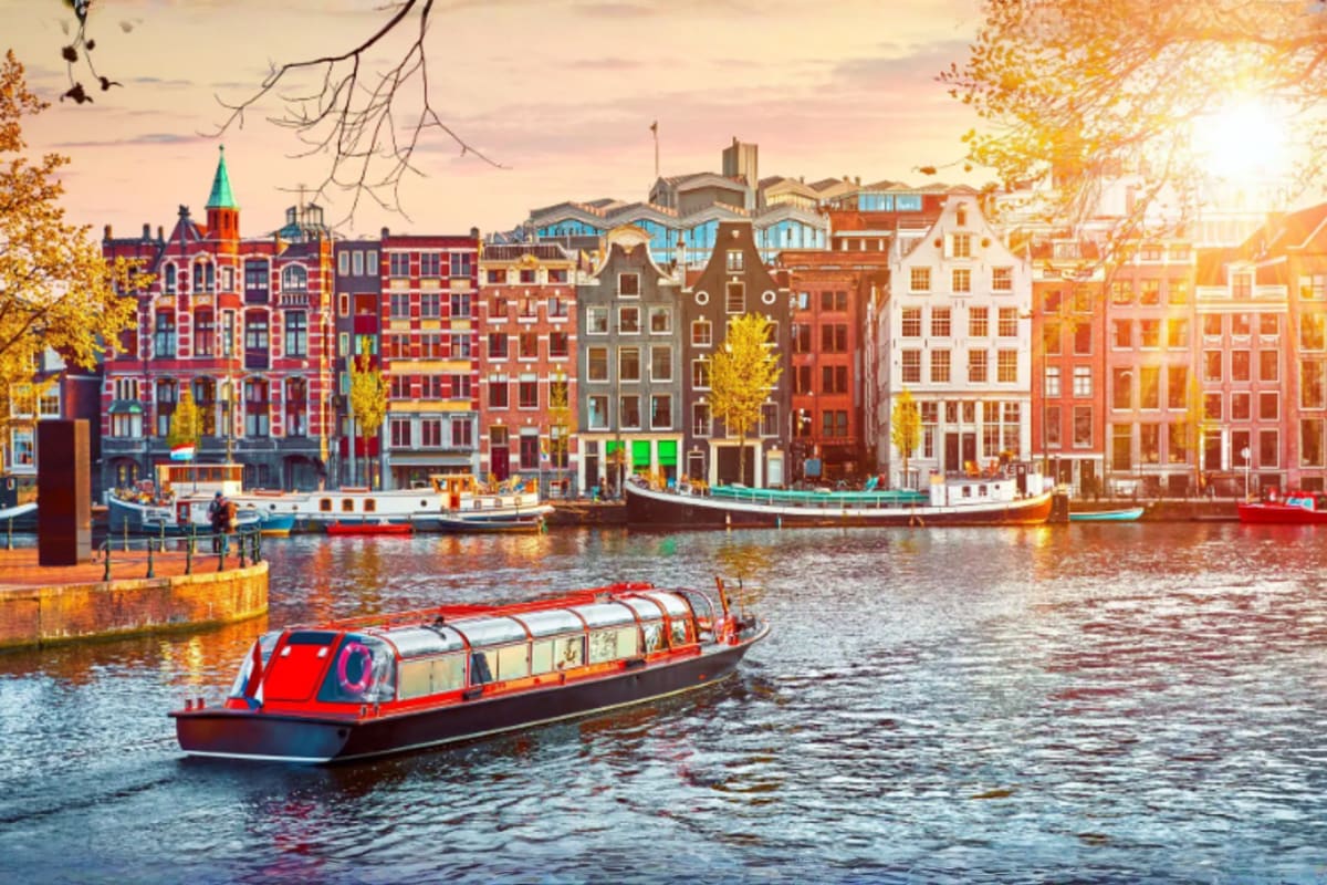 Red and white canal boat on Amsterdam waterway with historic colorful buildings and autumn foliage in background at sunset