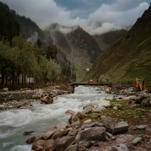 Mountain valley with rushing river, pine forests, and snow-capped peaks under cloudy sky in Kashmir