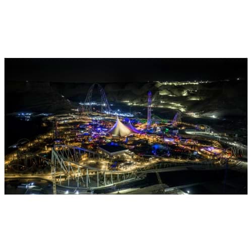 Aerial night view of Qiddiya theme park illuminated with blue and gold lights, featuring roller coasters and carnival attract