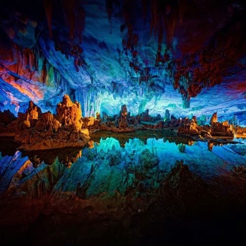 Illuminated stalactites and stalagmites in a cave with blue and orange lighting reflecting in underground lake water