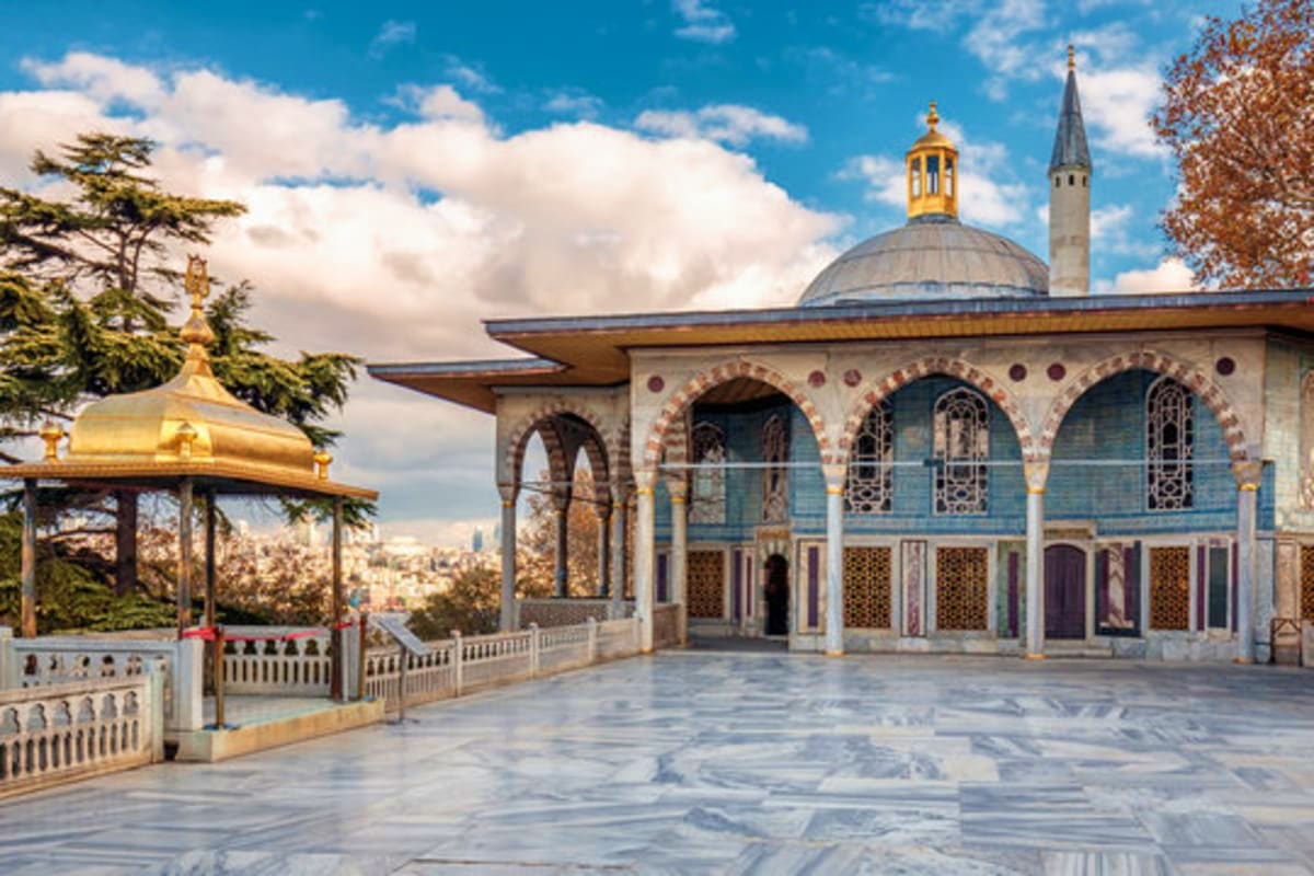 Ottoman-era mosque with blue tilework and golden dome, marble courtyard with arches, and city skyline in background under blu