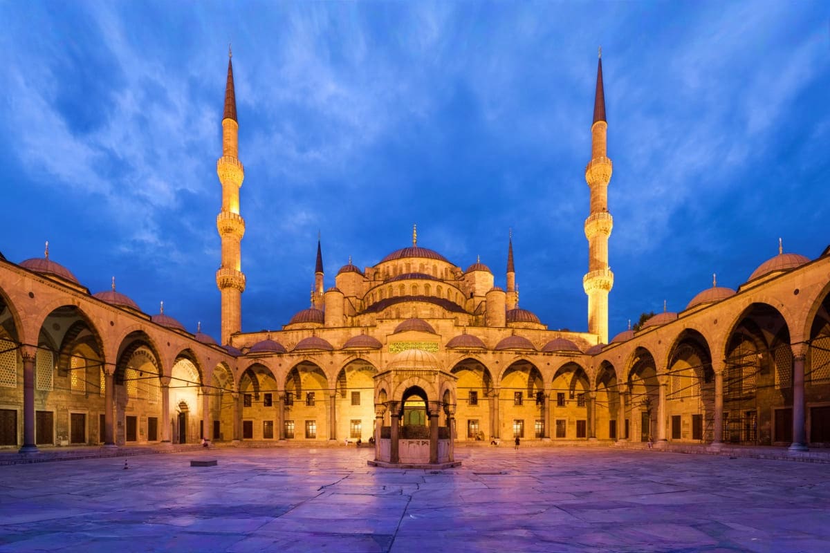 Blue Mosque in Istanbul at dusk with illuminated minarets and courtyard, showcasing Ottoman Islamic architecture