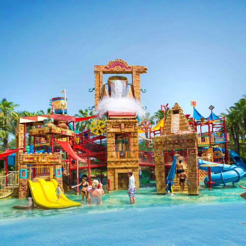 Colorful water park with large wooden-themed play structures, slides, and a massive bucket dumping water, surrounded by child