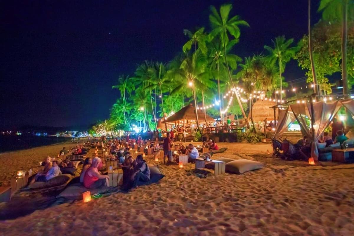 Night beach festival with crowds seated on sand, illuminated palm trees, beachfront tents, and ocean view under starry sky