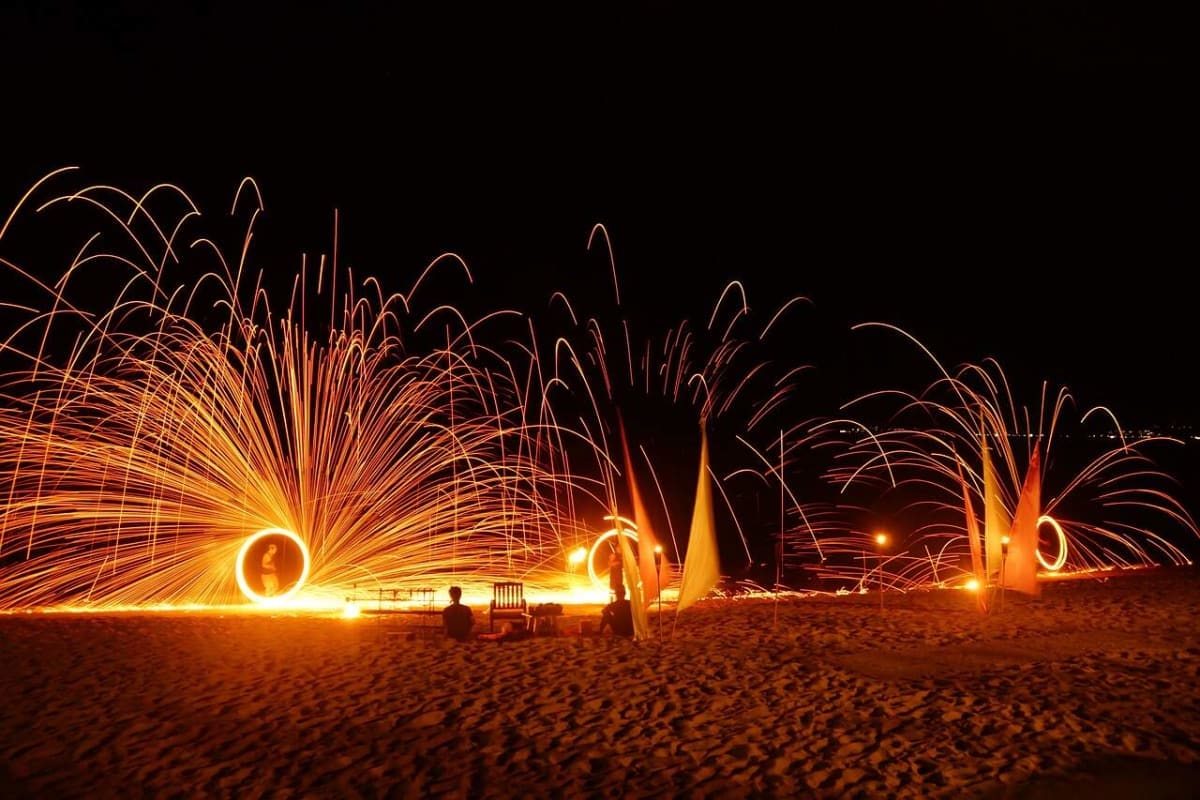 Long exposure photograph of steel wool fire spinning creating golden light trails on a beach at night with spectators