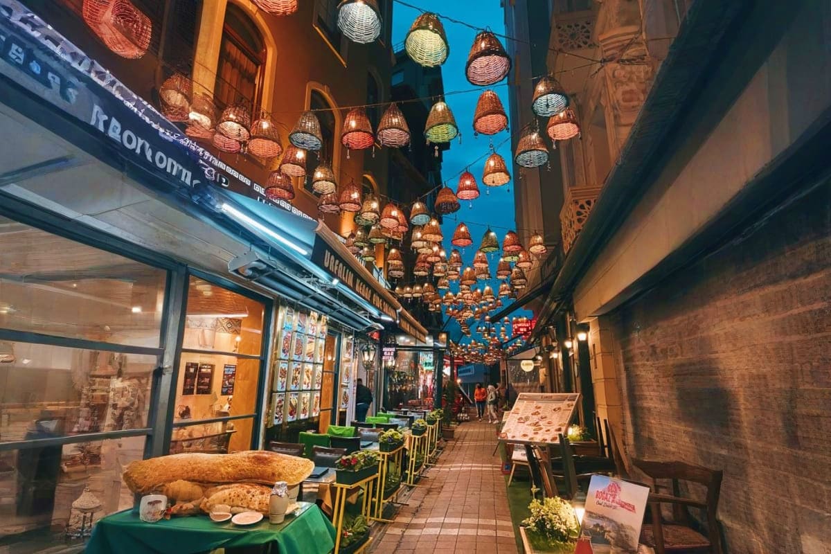 Narrow alleyway with illuminated decorative lanterns hanging overhead, shop windows with displays, and green chairs lining a 