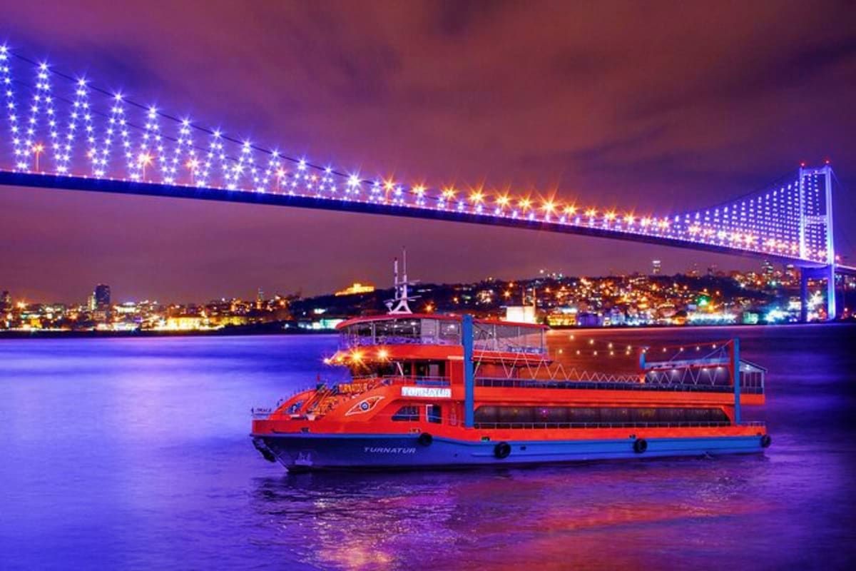 Red and blue passenger ferry boat on illuminated water at night under brightly lit suspension bridge in Istanbul