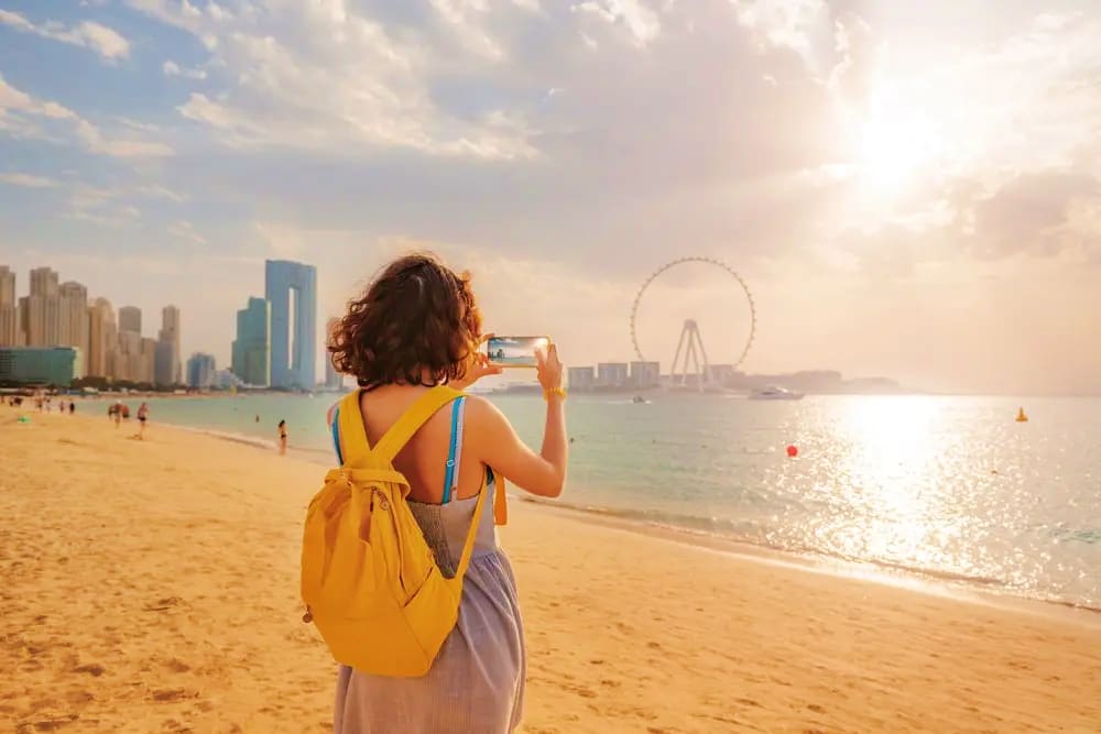 Woman photographing Dubai skyline and Ferris wheel from Jumeirah Beach at sunset