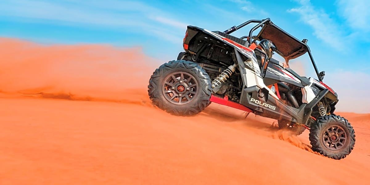 Polaris RZR off-road vehicle driving across orange sand dunes in Dubai desert under blue sky
