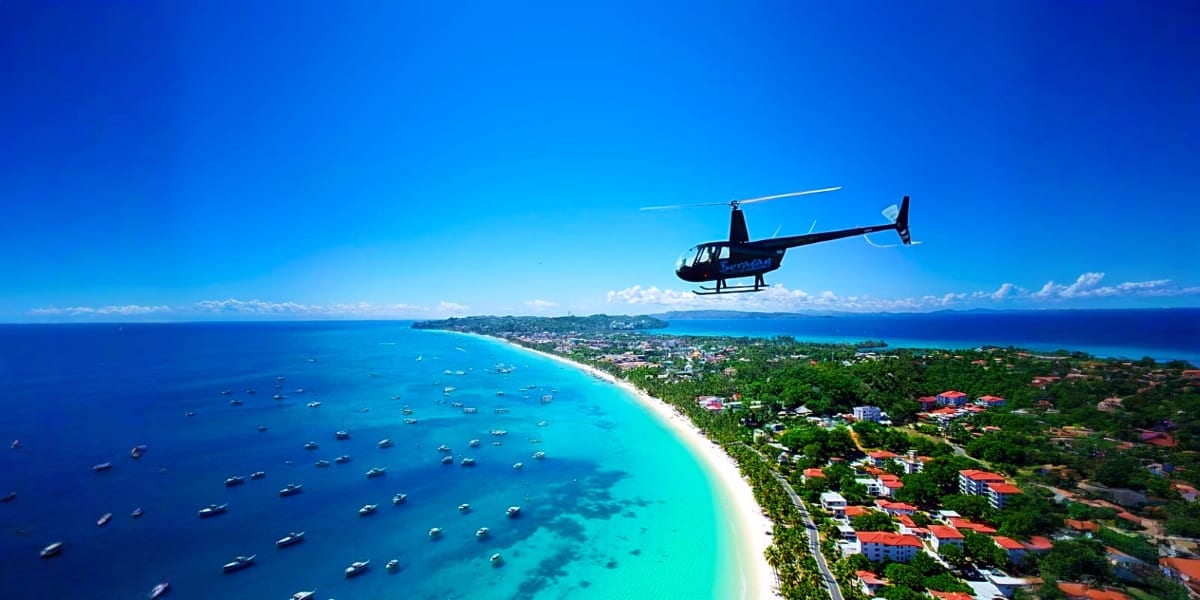Helicopter flying over Boracay Island's turquoise waters, white sand beach, and coastal village with boats anchored offshore