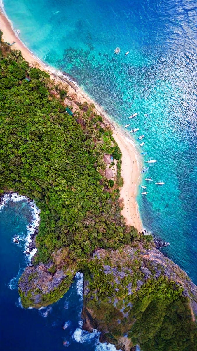 Aerial view of tropical island with sandy beach, turquoise waters, and anchored boats, surrounded by lush green forest and ro