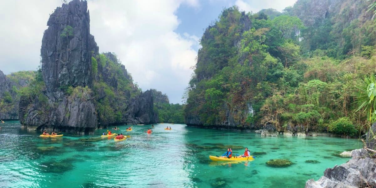Kayakers paddling in turquoise lagoon surrounded by tall limestone cliffs and lush vegetation in El Nido, Palawan