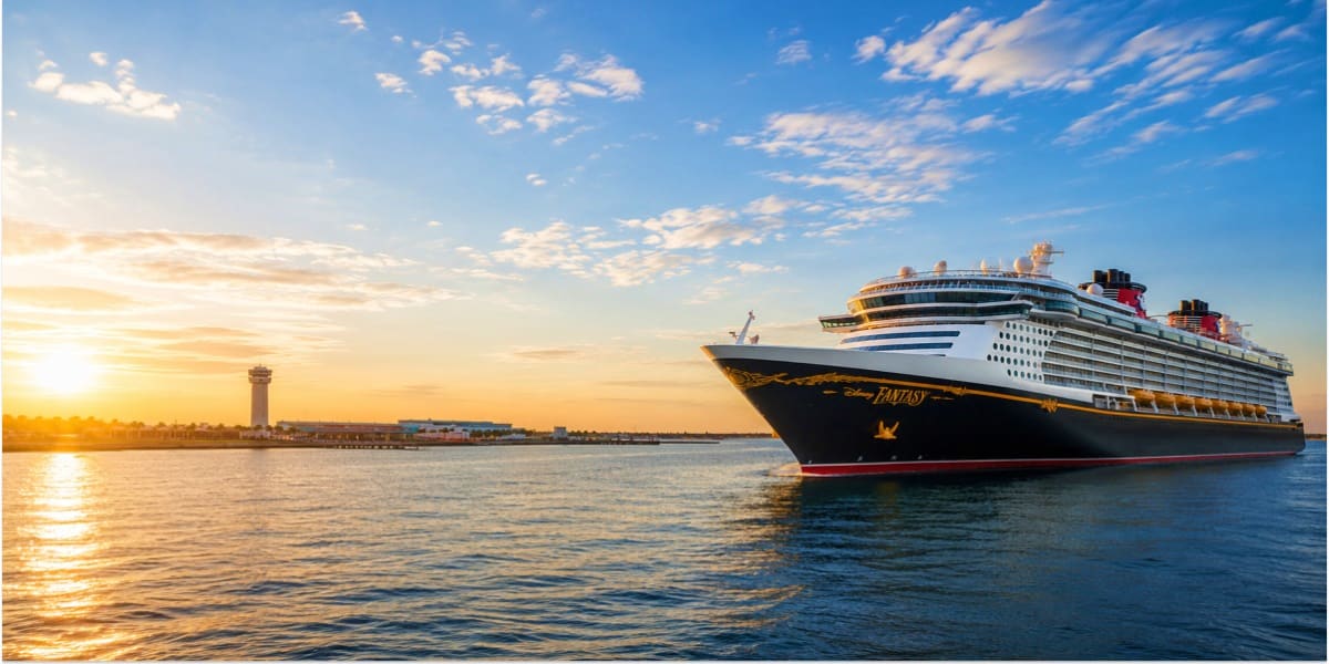Large cruise ship Disney Fantasy sailing at sunrise with lighthouse and port terminal visible on the horizon