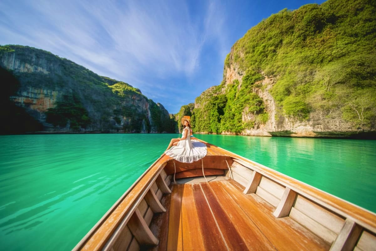 04 Island Tour on Speed Boat Woman in white dress sitting on wooden speedboat bow in turquoise water surrounded by tall green limestone cliffs