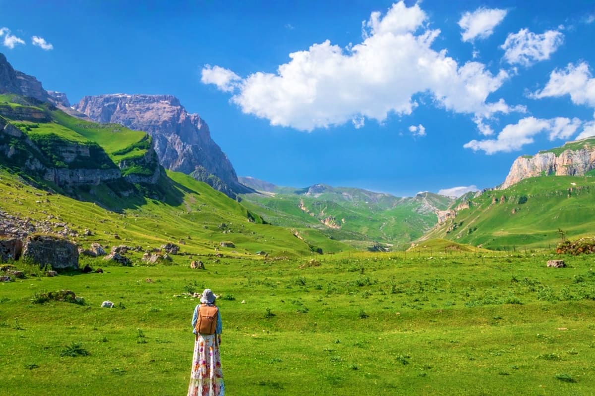 Hiker with backpack standing in lush green alpine meadow surrounded by dramatic rocky mountain peaks under blue sky with whit