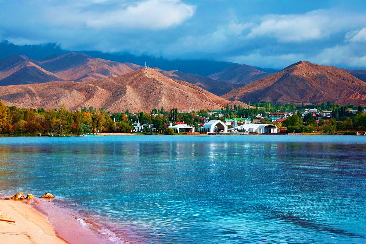 Scenic view of Issyk Kul Lake with turquoise water, sandy beach in foreground, tree-lined shoreline with buildings, and barre