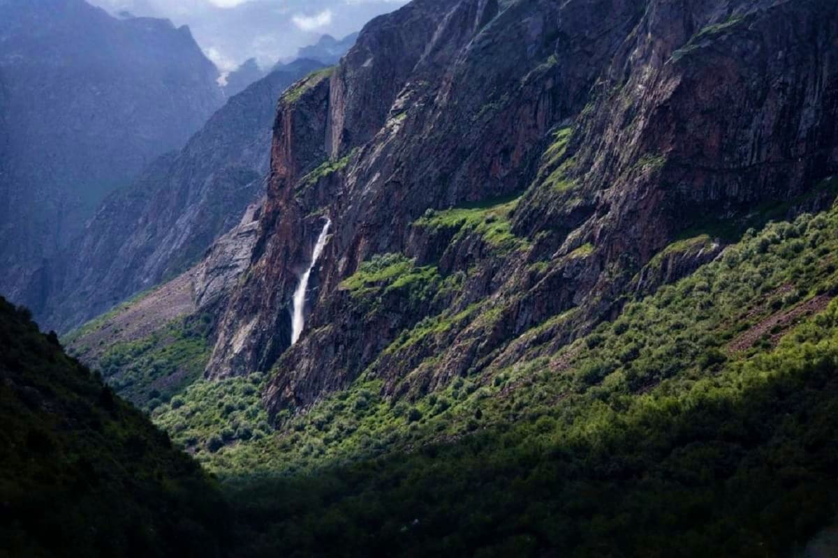 Waterfall cascading down steep rocky cliff faces in a forested mountain valley with lush green vegetation