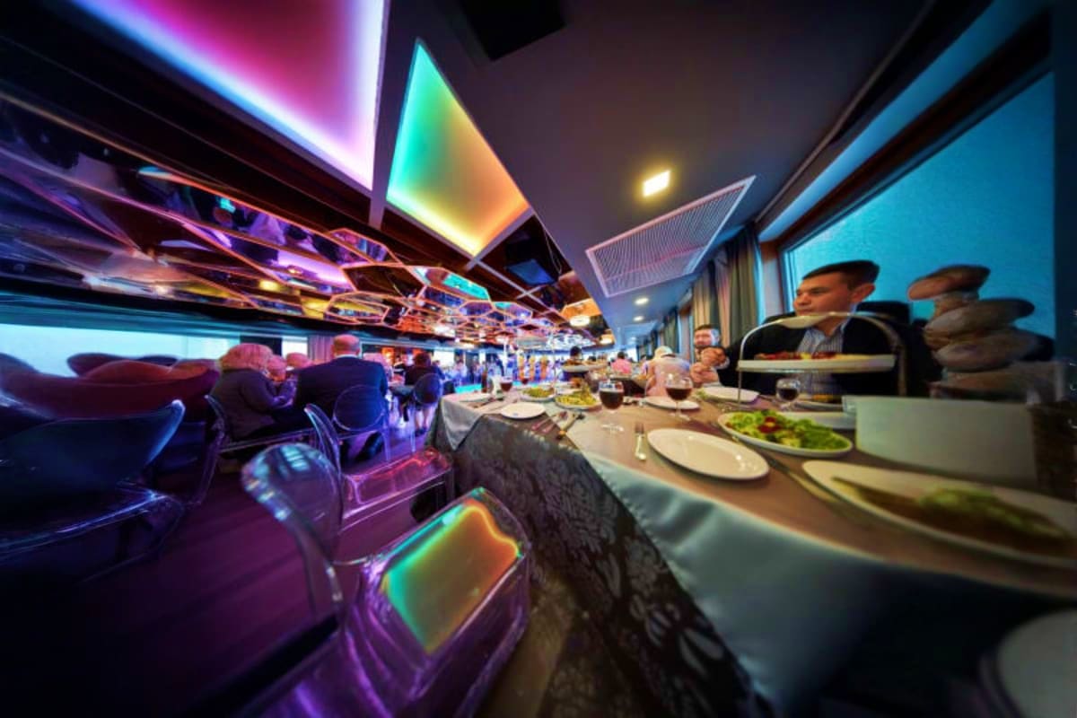 All star cruise Luxury coach interior with dining area, passengers seated at tables with meals, colorful neon lighting on ceiling, large wind