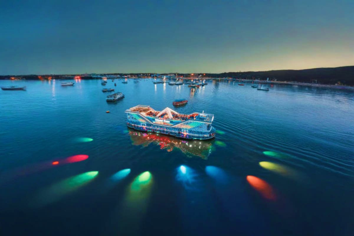 All star cruise Aerial view of colorful lit cruise ship anchored in calm blue bay with fishing boats and coastal town at dusk