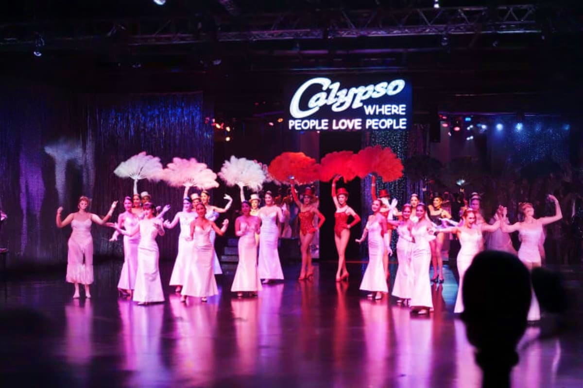 Cabaret performers in pink and red dresses holding oversized feathered umbrellas on stage at Calypso Bangkok nightclub with i