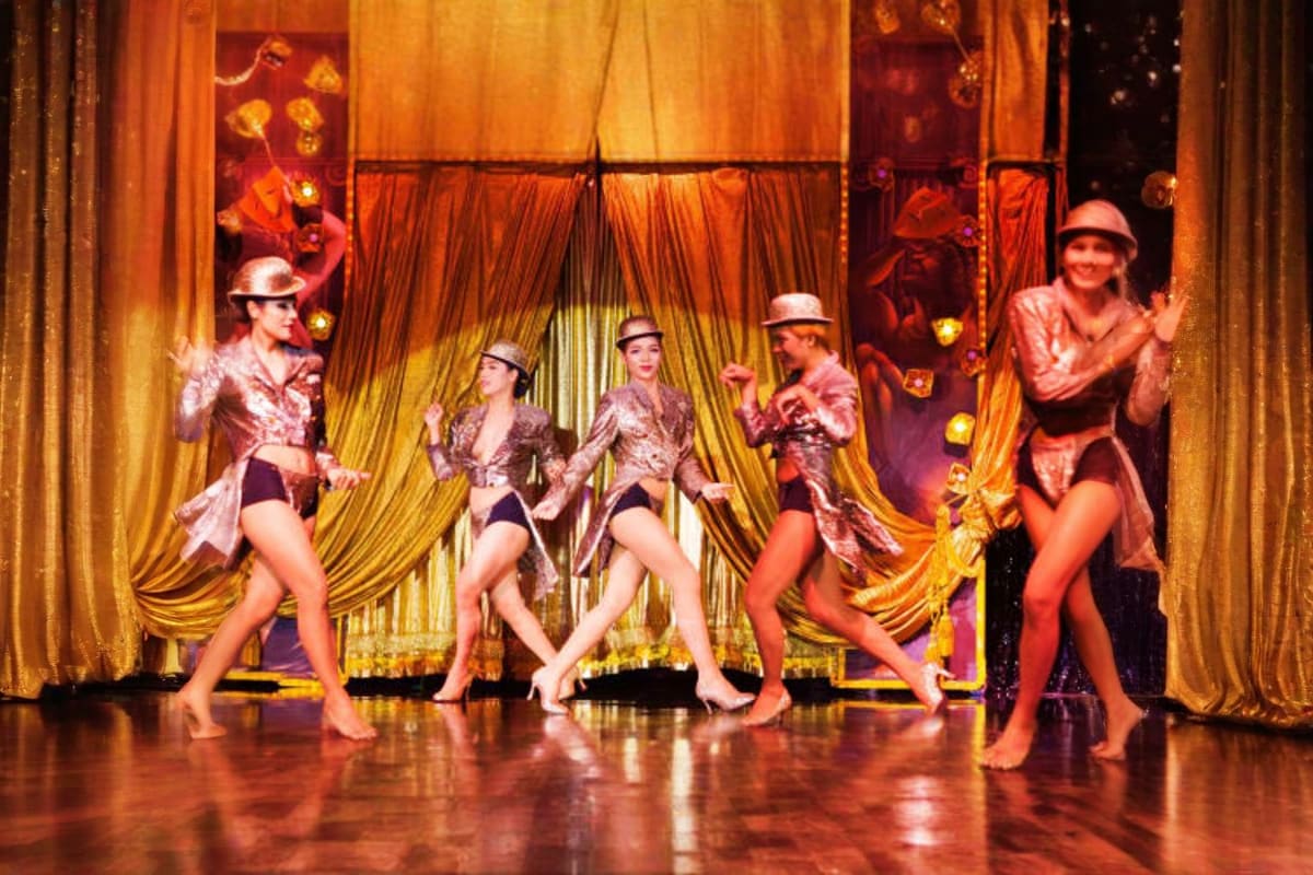 Cabaret performers in sequined outfits and fedoras dancing on stage with gold curtains and theatrical lighting at Calypso Ban