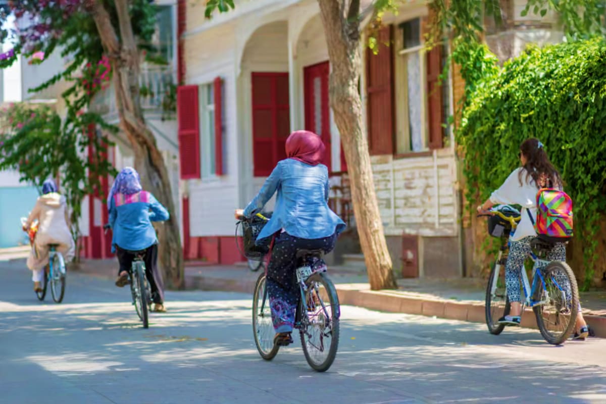 Children riding bicycles on a tree-lined street with red and white traditional buildings in a Mediterranean village setting