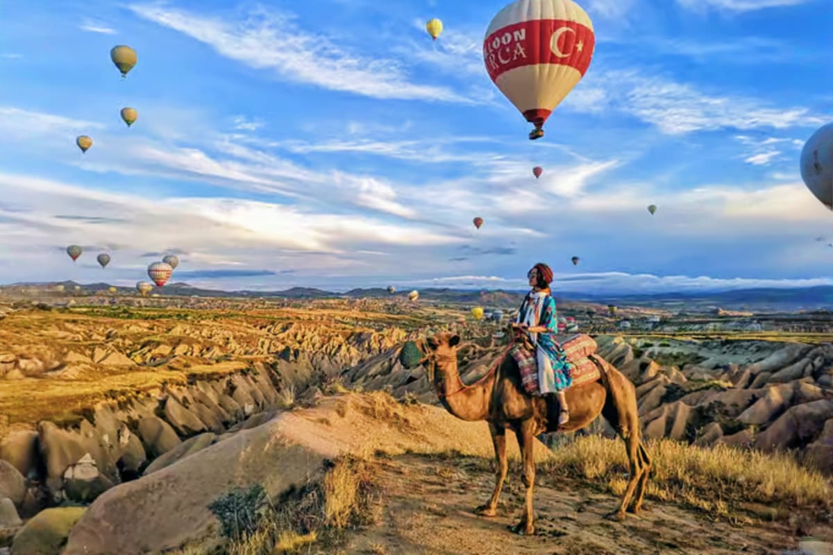 Person riding camel in Cappadocia's rocky landscape with hot air balloons floating in blue sky above
