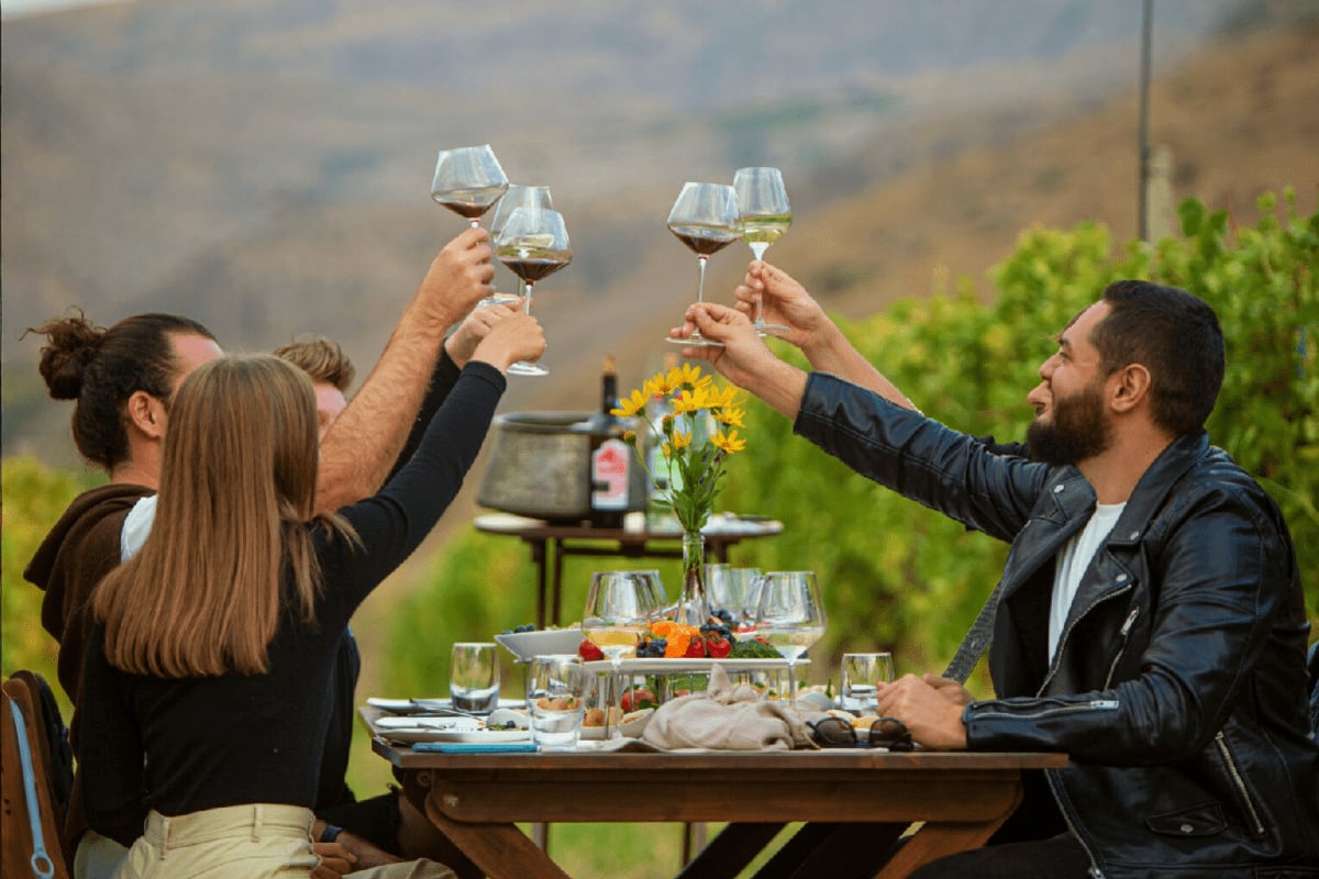 Group of four people toasting wine glasses at an outdoor dining table with vineyard hillsides and green grapevines in the bac