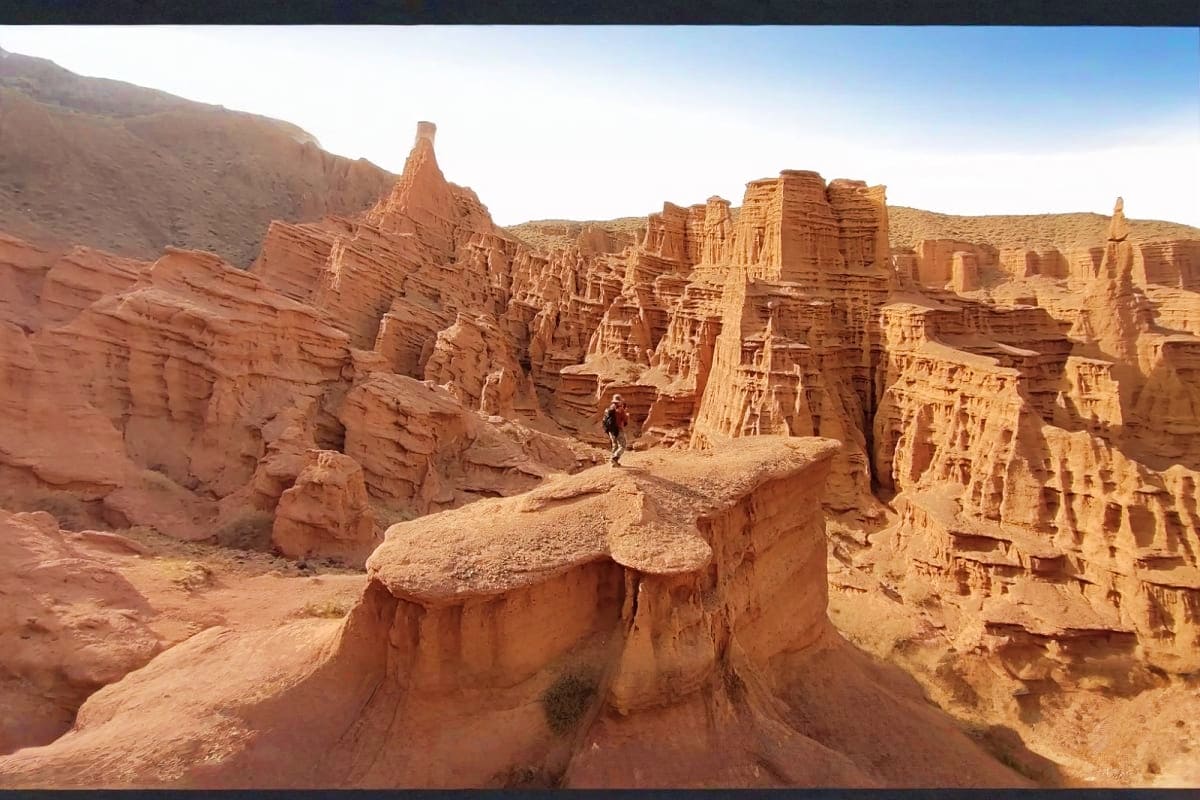 Ancient carved rock formations and structures in a desert canyon landscape with a hiker on the trail