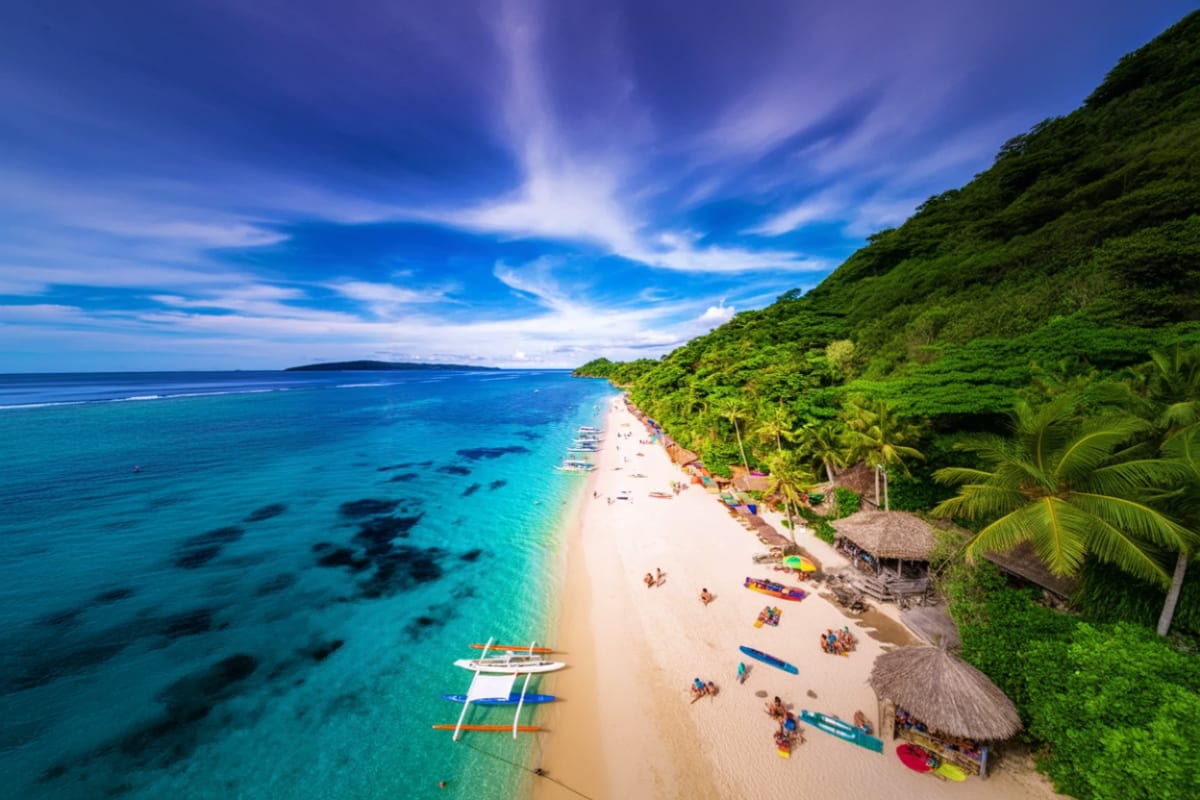 Aerial view of a pristine white sand beach with turquoise waters, traditional boats, palm trees, and jungle cliffs on Boracay