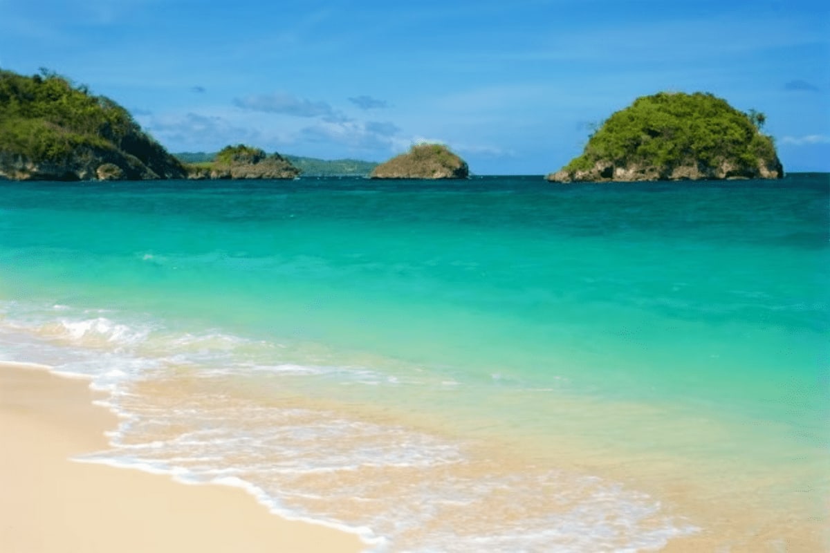 Turquoise beach with white sand and gentle waves, small forested islands visible across clear blue water under sunny sky