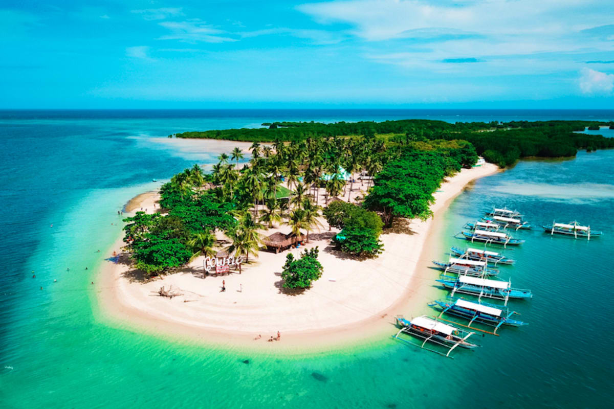Aerial view of a tropical island with white sand beach, palm trees, thatched-roof structures, and traditional Filipino boats 