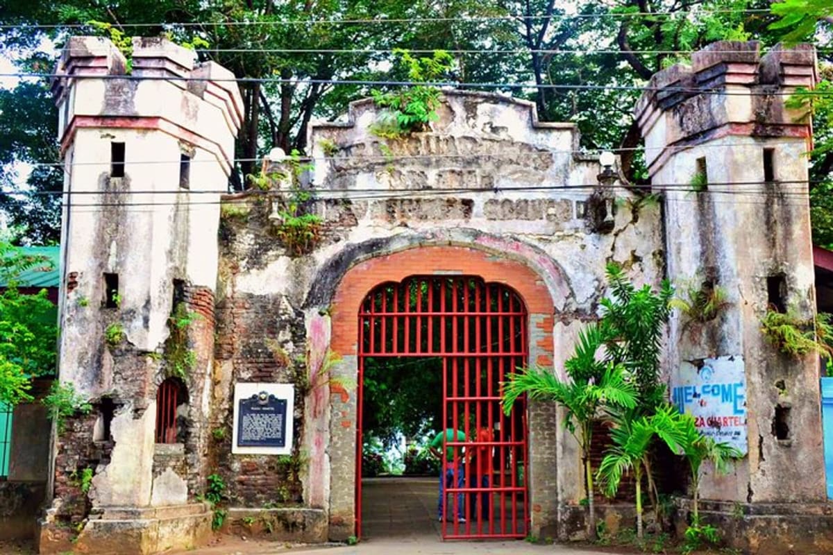 Historic colonial fort with weathered white walls, red barred gate, twin towers, and surrounding green vegetation