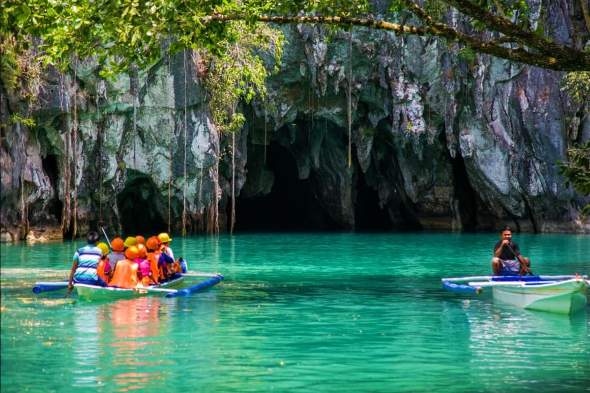 Tourists in orange life jackets on boats in turquoise waters inside a limestone cave with towering rock formations and lush g