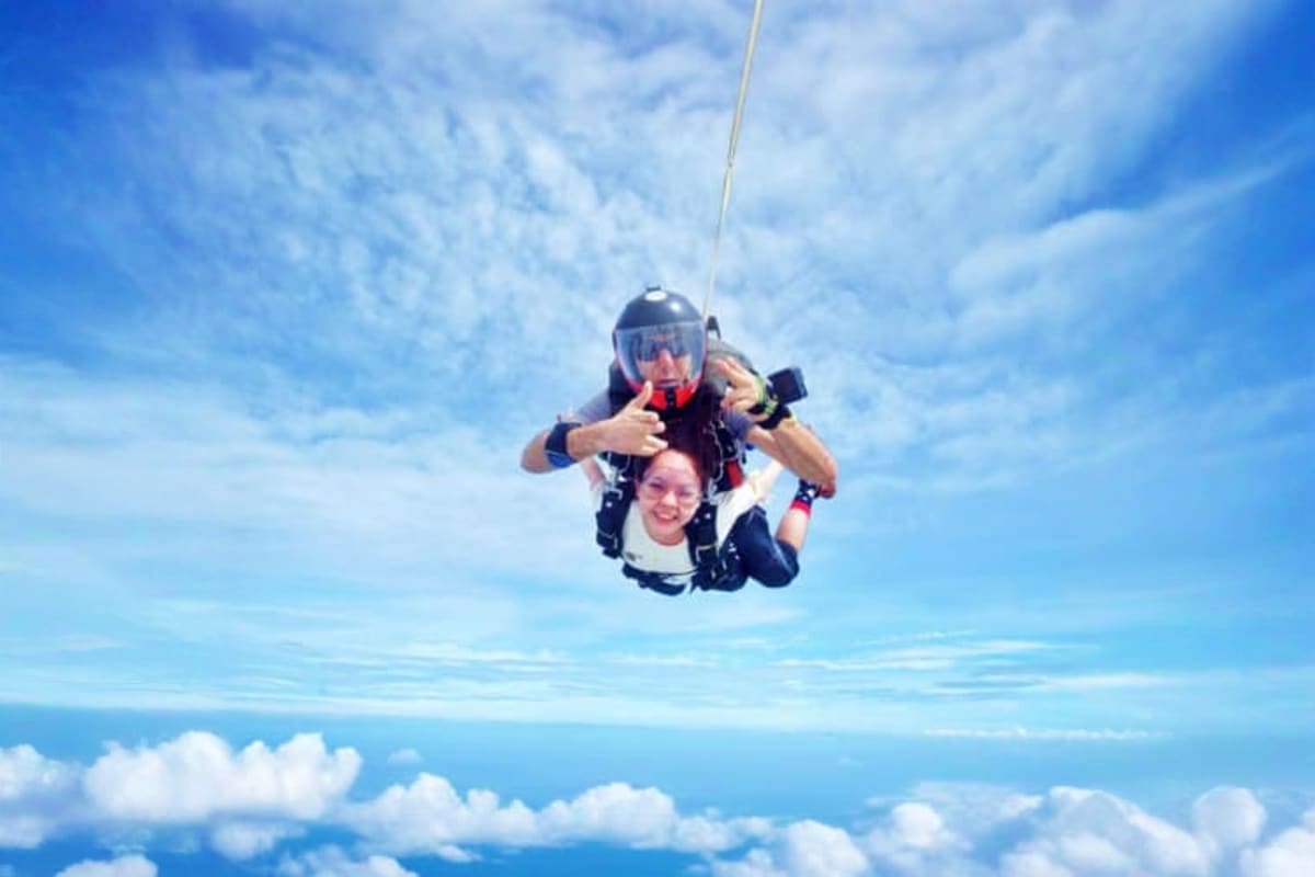 Tandem skydiving instructor and student descending through blue sky with parachute deployed above clouds