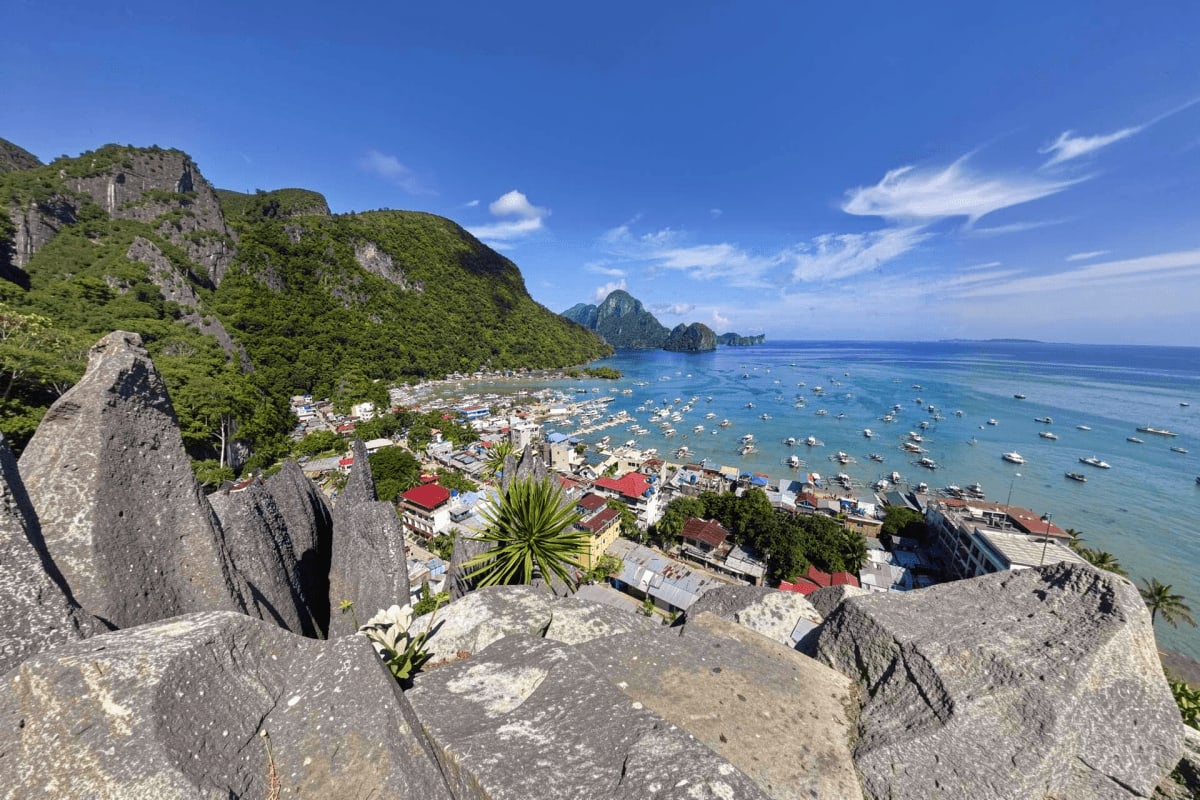 Elevated view of El Nido coastal town with limestone cliffs, turquoise waters, and numerous boats anchored in the bay under c