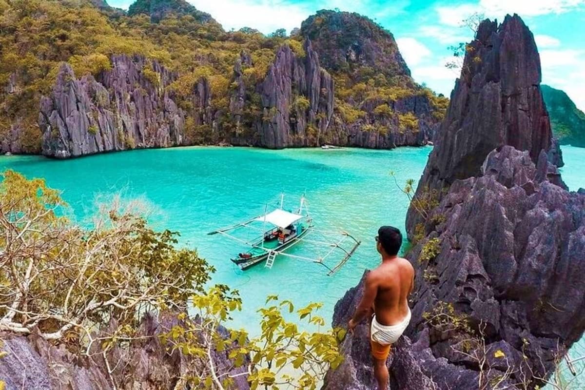 Man standing on limestone cliff overlooking turquoise lagoon with traditional boats and towering rock formations in El Nido, 