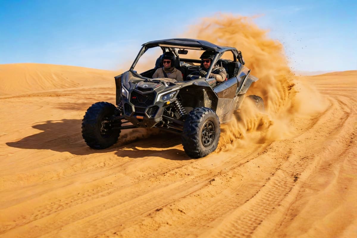 Two riders in a Can-Am Maverick buggy racing through golden desert sand, creating a large dust cloud behind them under blue s