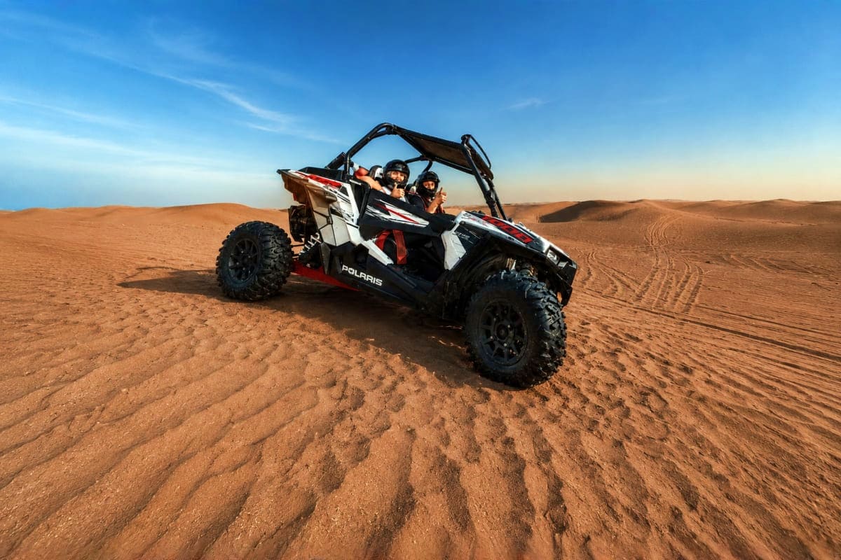 Two people riding a red and white Polaris RZR side-by-side through golden desert sand dunes under blue sky
