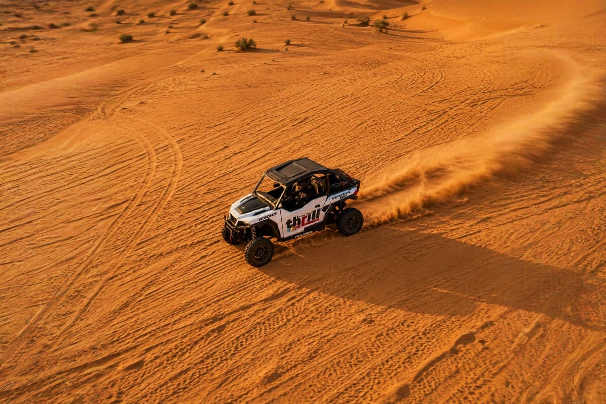 White and black Polaris General UTV kicking up sand dust while driving across golden desert dunes with sparse vegetation