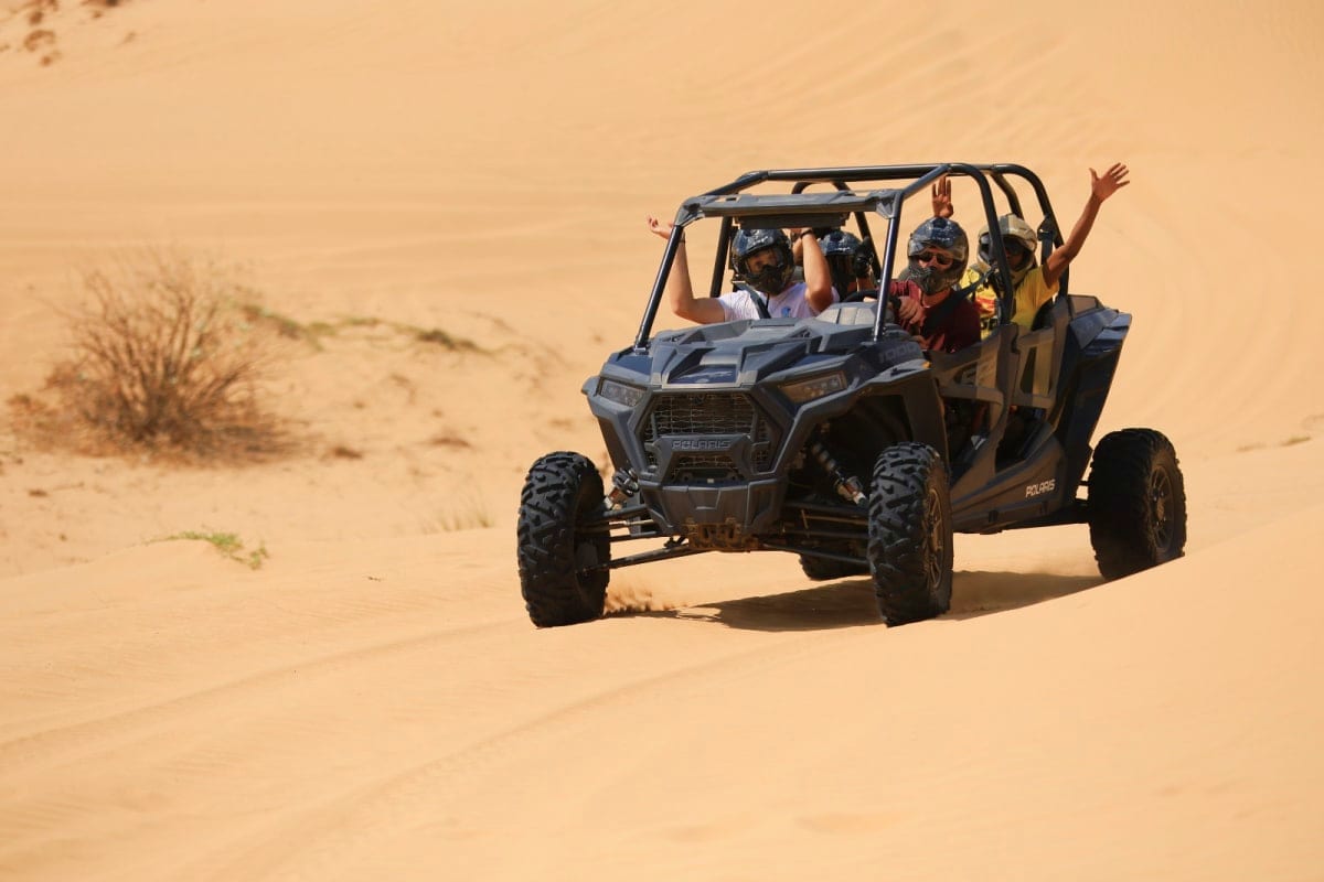 Group of four people riding in a Polaris RZR 1000cc four-seater buggy across sandy desert dunes