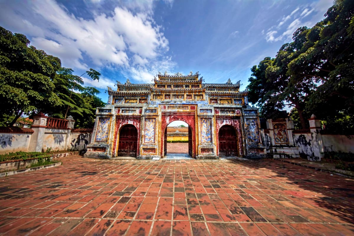 Hue City tour Traditional Vietnamese gateway with ornate golden roof and red doors at the Citadel in Hue, framed by lush trees and brick co