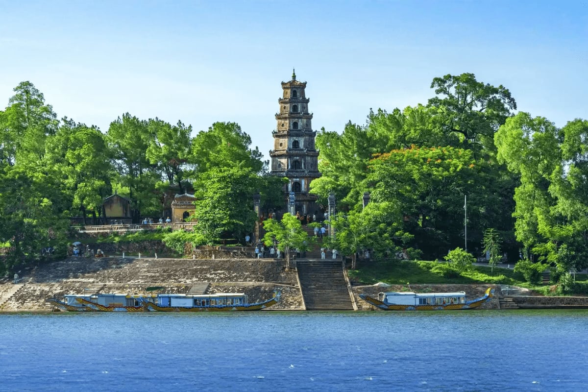 Hue City tour Thien Mu Pagoda tower rising above lush green trees on the Perfume River in Hue, Vietnam, with traditional blue boats moored
